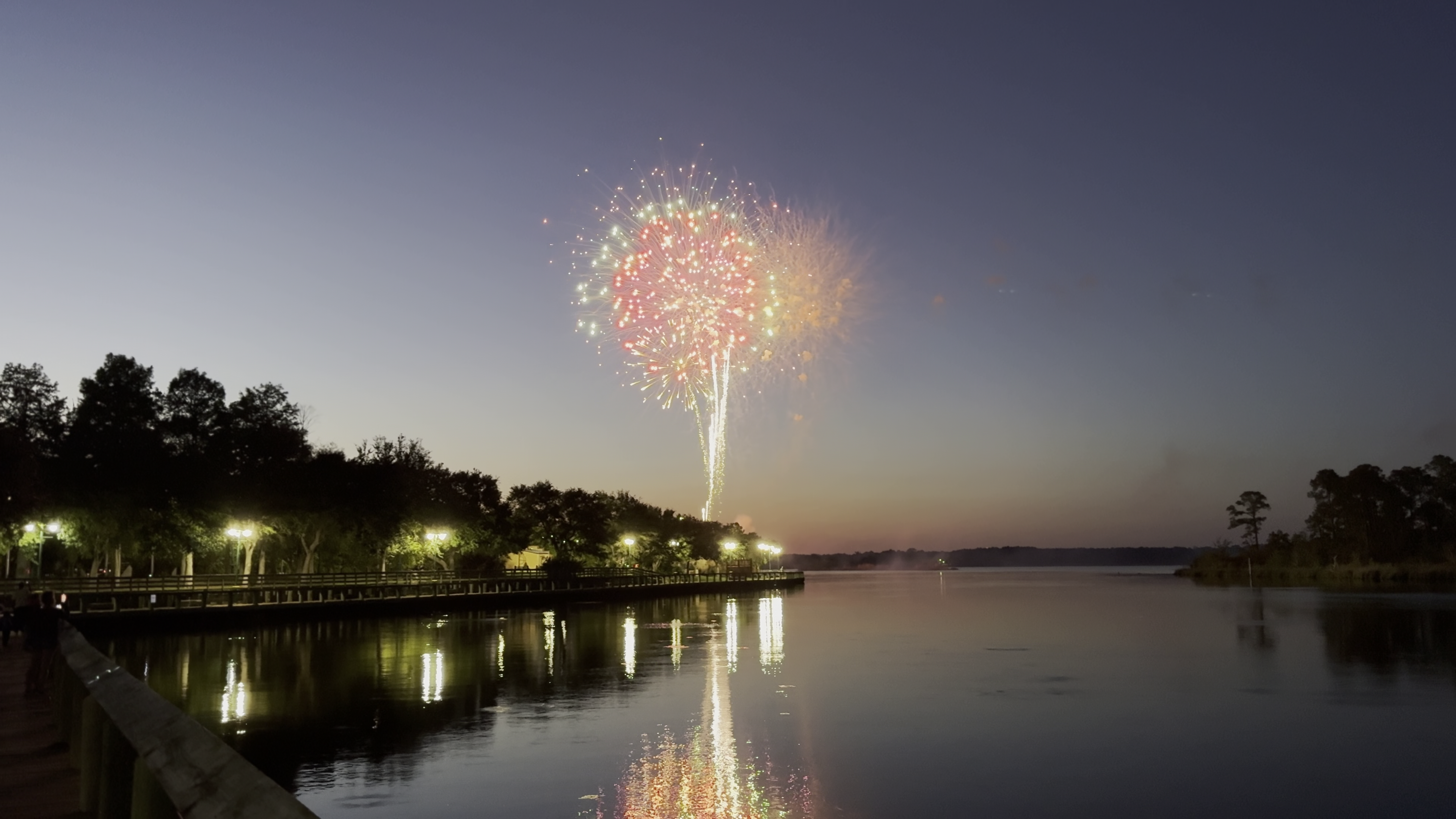 Fireworks burst over the Pascagoula River at twilight during River Jamboree 2026, reflected in the water from the Downtown Riverfront pier